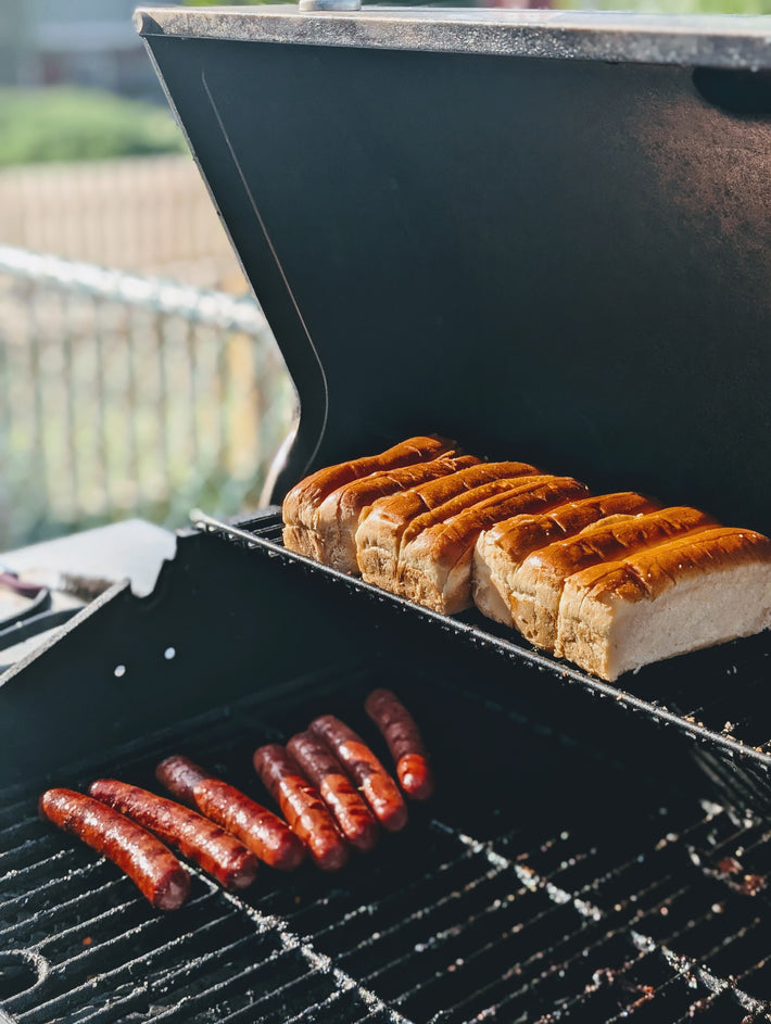 Toasted hot dog buns on the upper grill rack with several browned sausages cooking on the lower grate.