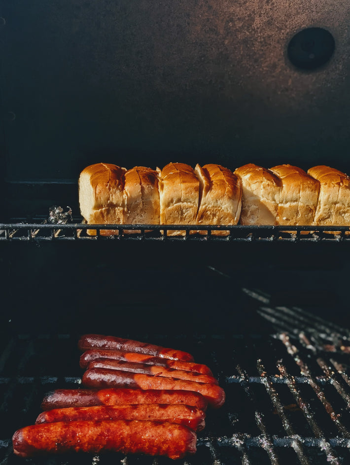Hot dogs browning on a grill grate with split sandwich rolls toasting on the upper rack