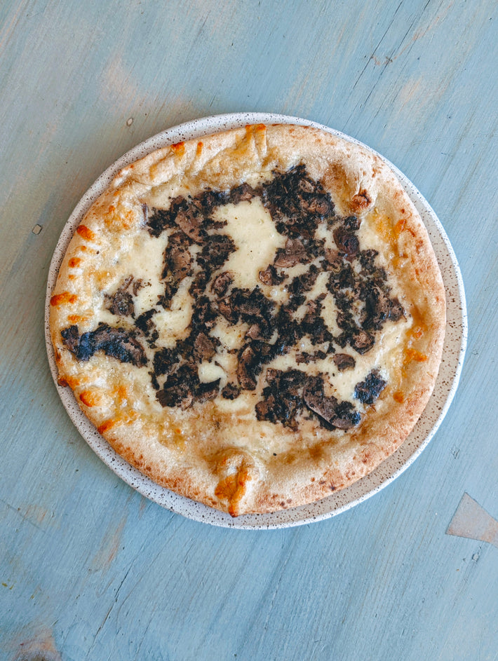 Round pizza on a speckled plate with melted cheese and scattered dark mushroom-like pieces, on a blue wooden table.