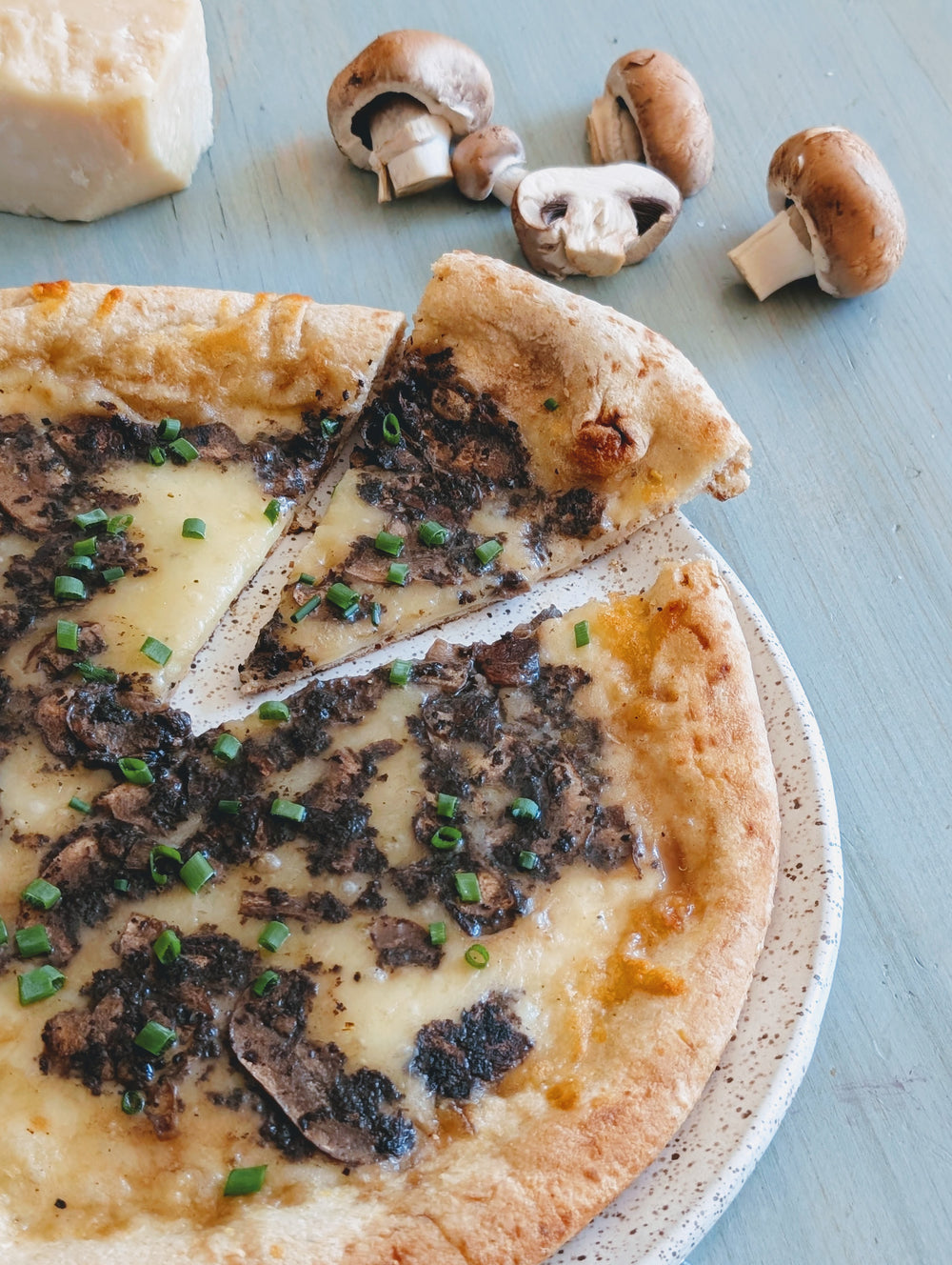 Sliced mushroom and chive pizza with melted cheese on a speckled plate, cremini mushrooms and a cheese wedge nearby