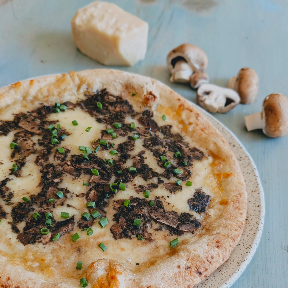Wood-fired pizza with melted cheese, sliced mushrooms and chopped chives on a speckled plate; mushrooms and a cheese wedge behind it.