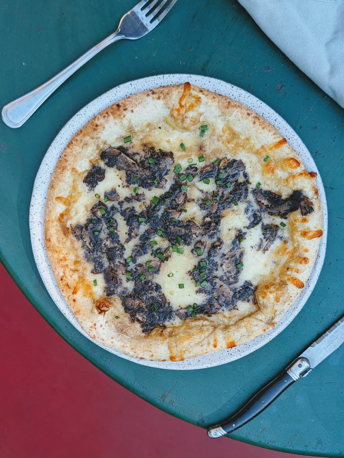 Round pizza with melted mozzarella, dark truffle-mushroom topping and chopped chives on a plate, utensils on green table.