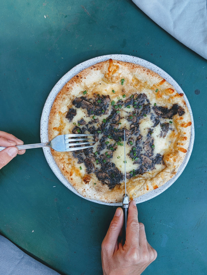 Hands cutting a mushroom-and-chive pizza on a speckled plate over a teal table.