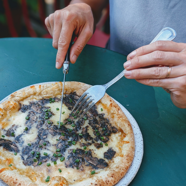 Hands using a fork and knife to cut a mushroom-and-chive pizza with melted cheese on a speckled plate at a green table.