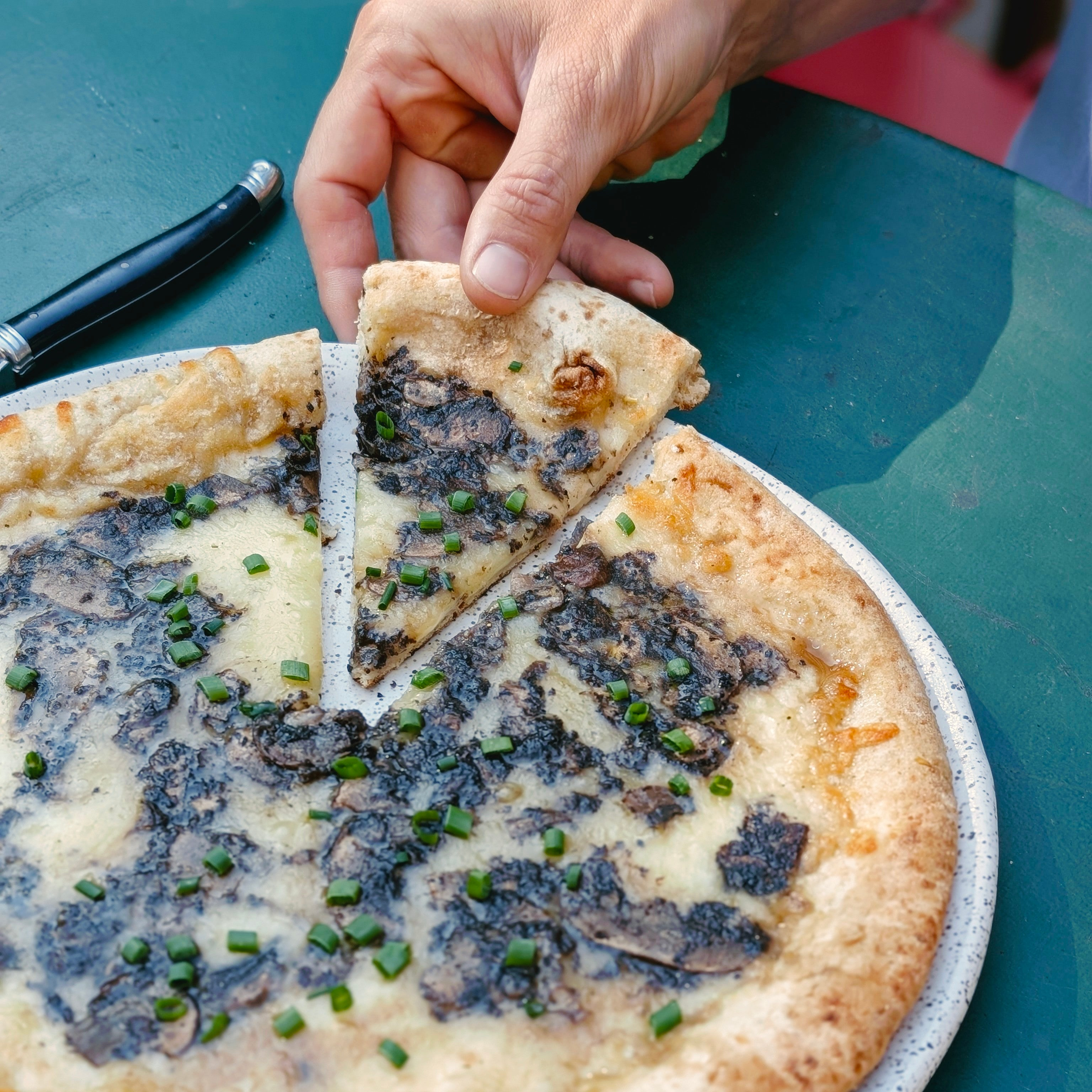 Hand lifting a slice of truffle pizza topped with shaved black truffle and chopped chives from a round plate.