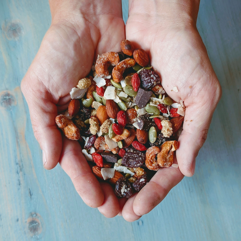 Cupped hands holding trail mix: almonds, cashews, pumpkin seeds, dried cranberries, coconut flakes and chocolate.