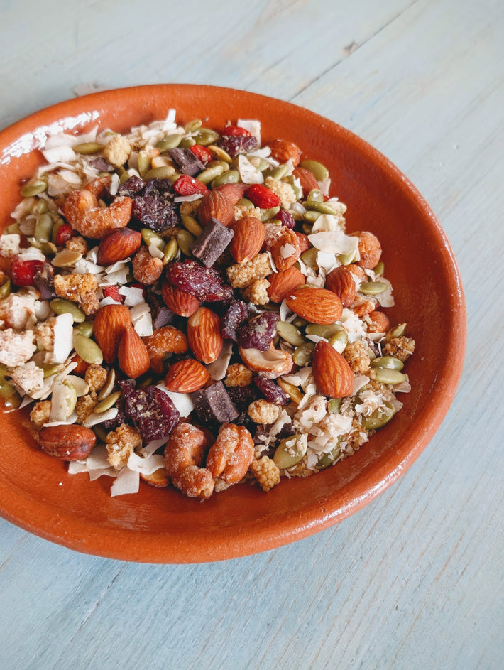 Terracotta bowl of trail mix with almonds, pepitas, dried cranberries, coconut flakes and chocolate chunks on blue wood.