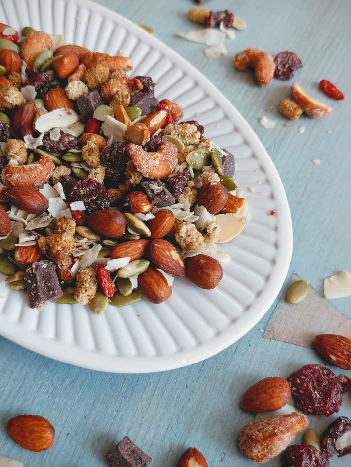 White plate with mixed trail mix: almonds, cashews, pumpkin seeds, dried cranberries, coconut flakes and chocolate chunks on blue table