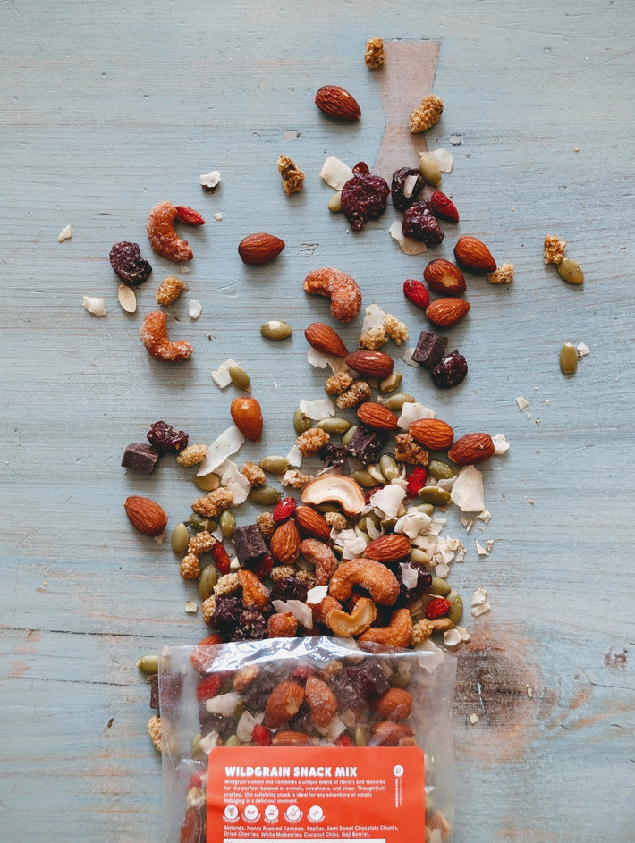 Nuts, seeds, dried fruit and chocolate spilling from a clear pouch labeled WILDGRAIN SNACK MIX on blue wood.