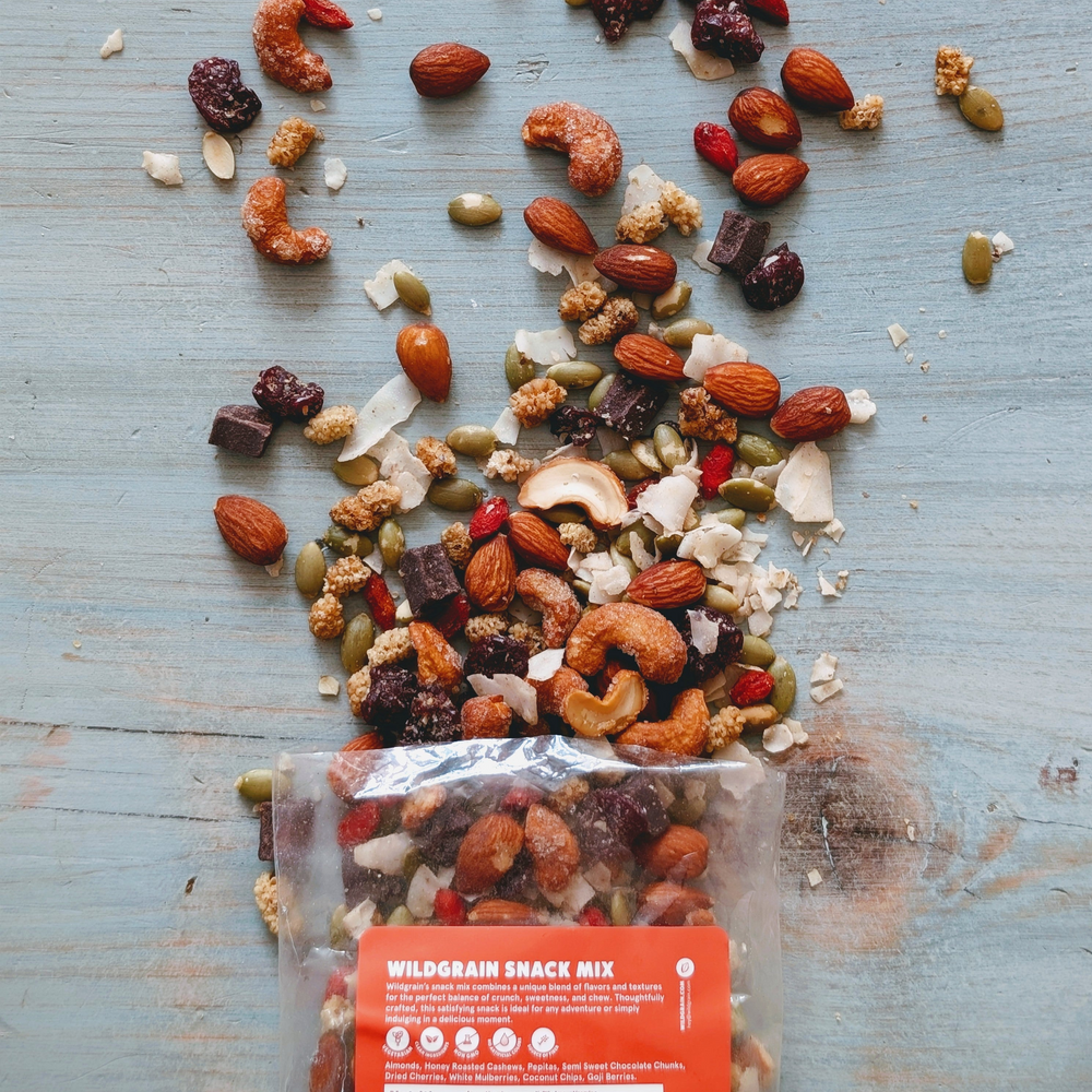 Bag labeled WILDGRAIN SNACK MIX with assorted cashews, almonds, seeds and dried fruits spilled on blue wooden surface