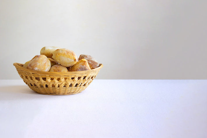 Woven basket of crusty bread rolls on a white table with plain light-gray background
