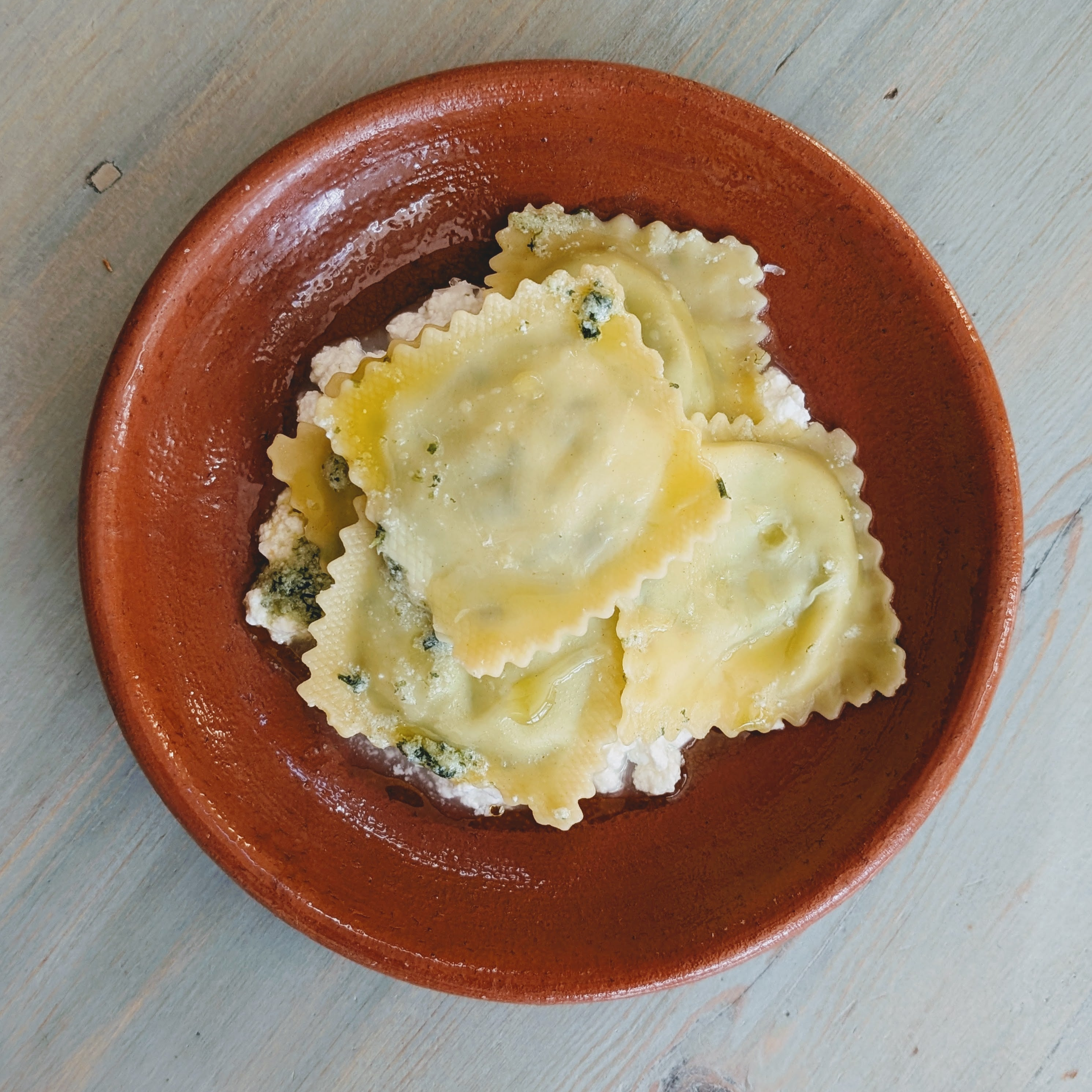 Three square ravioli on ricotta with olive oil in a brown ceramic bowl on a pale wood table.