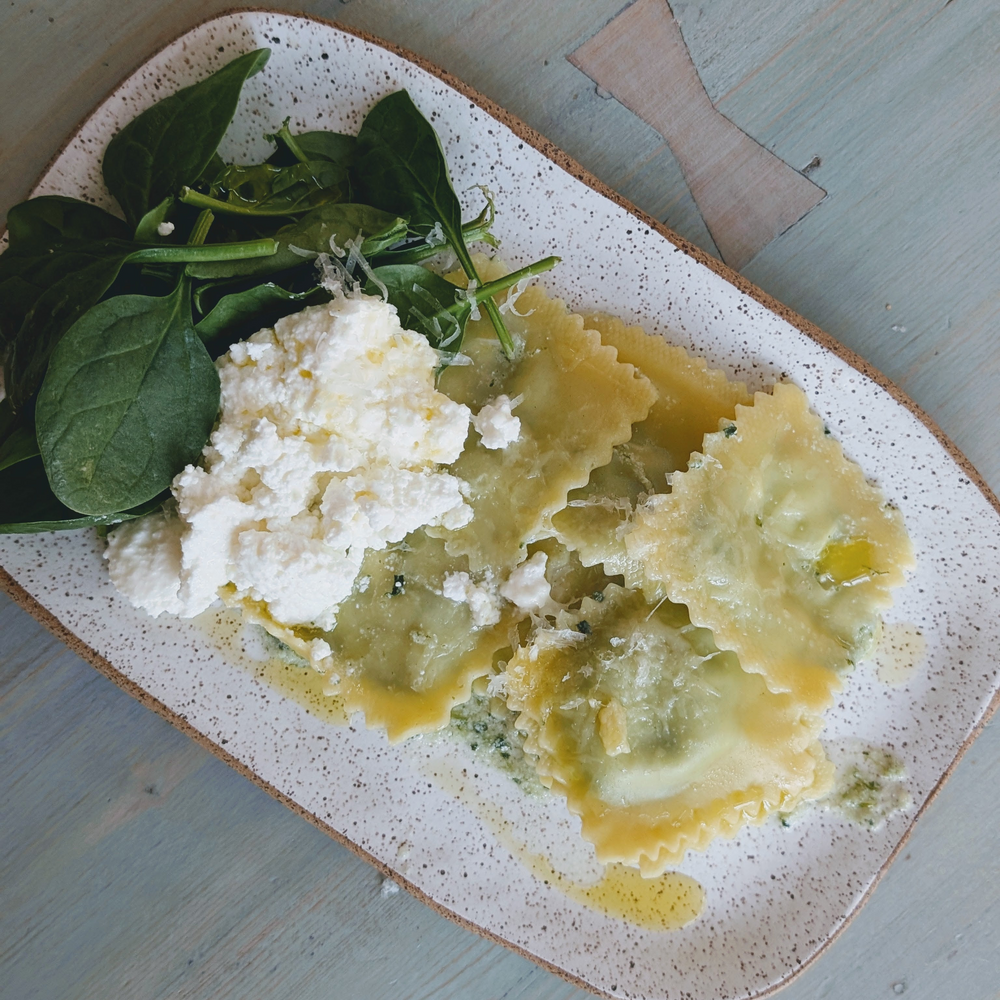 Top-down view of spinach ricotta ravioli on a white plate with ricotta crumbles and spinach garnish on wood