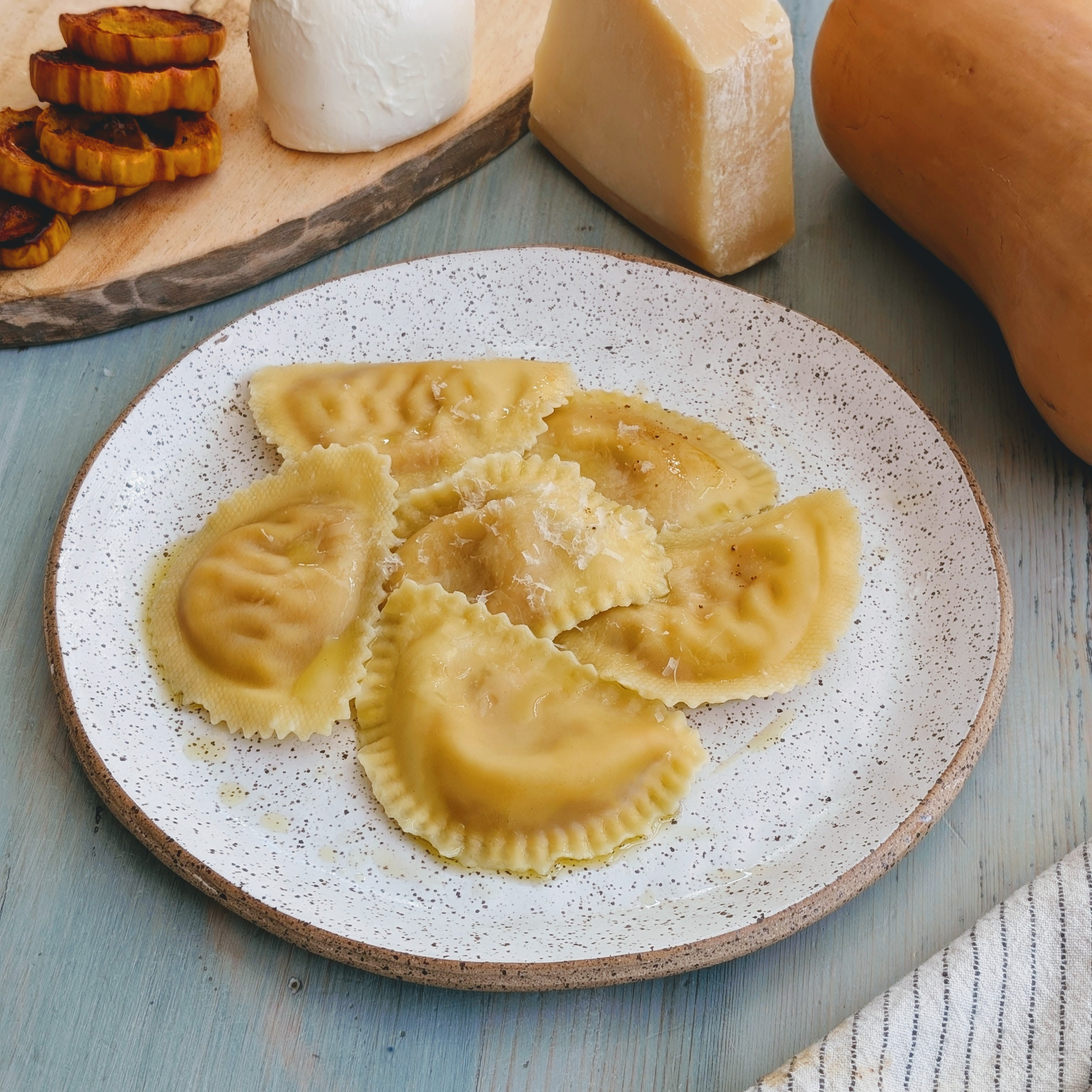 Ravioli drizzled with butter and grated cheese on a speckled plate, with a cheese wedge and roasted squash in the background