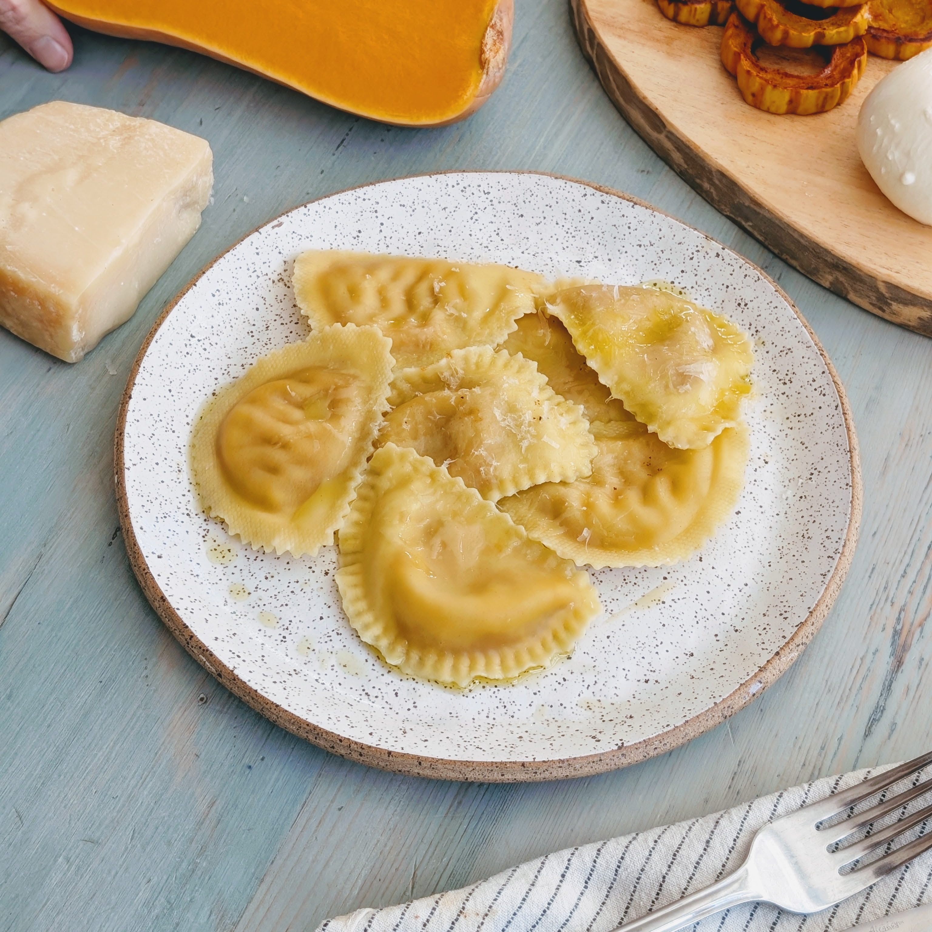 Plate of pumpkin ravioli drizzled with olive oil, with Parmesan wedge and roasted squash on a blue wooden table.