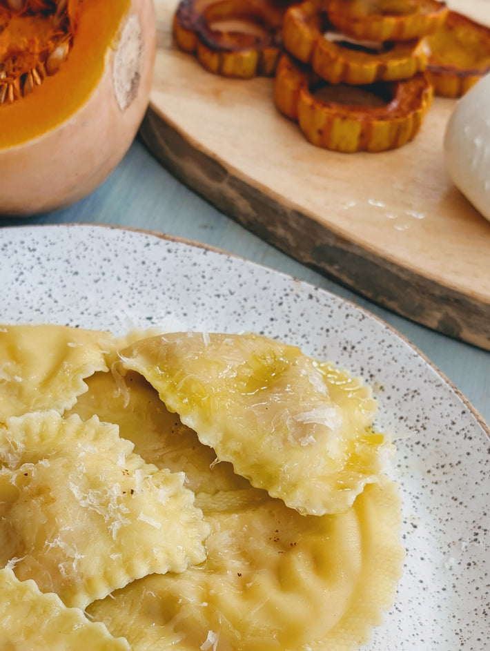 Butternut squash ravioli on a speckled plate, with a piece cut open, next to roasted squash slices and cheese on a wooden board.