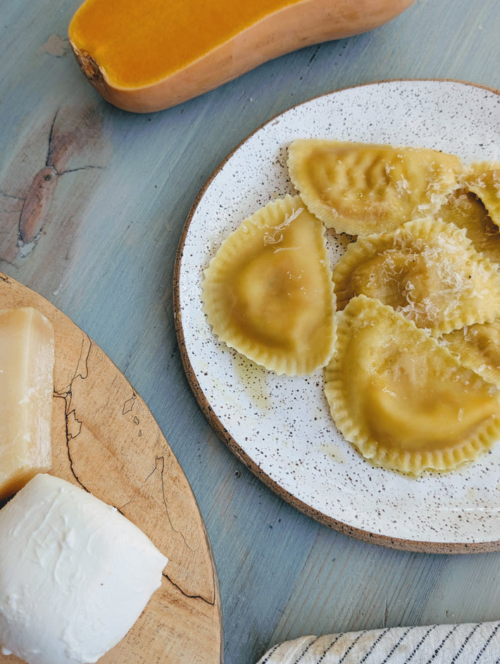 Butternut squash ravioli on a speckled plate with grated cheese, next to a butternut squash and cheese on a wooden board.