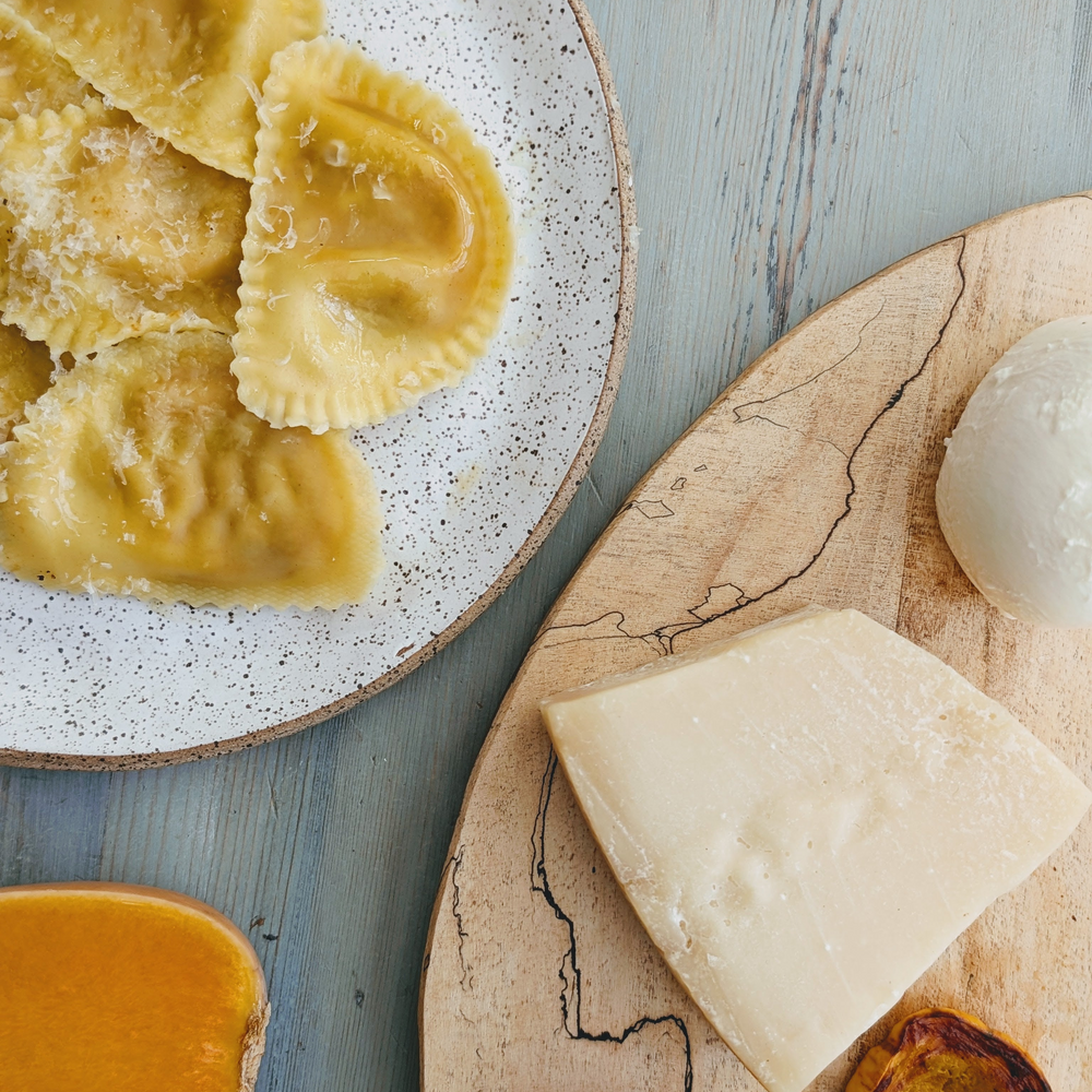 Ravioli on a speckled plate with a wedge of hard cheese and a ball of fresh mozzarella on a wooden board.