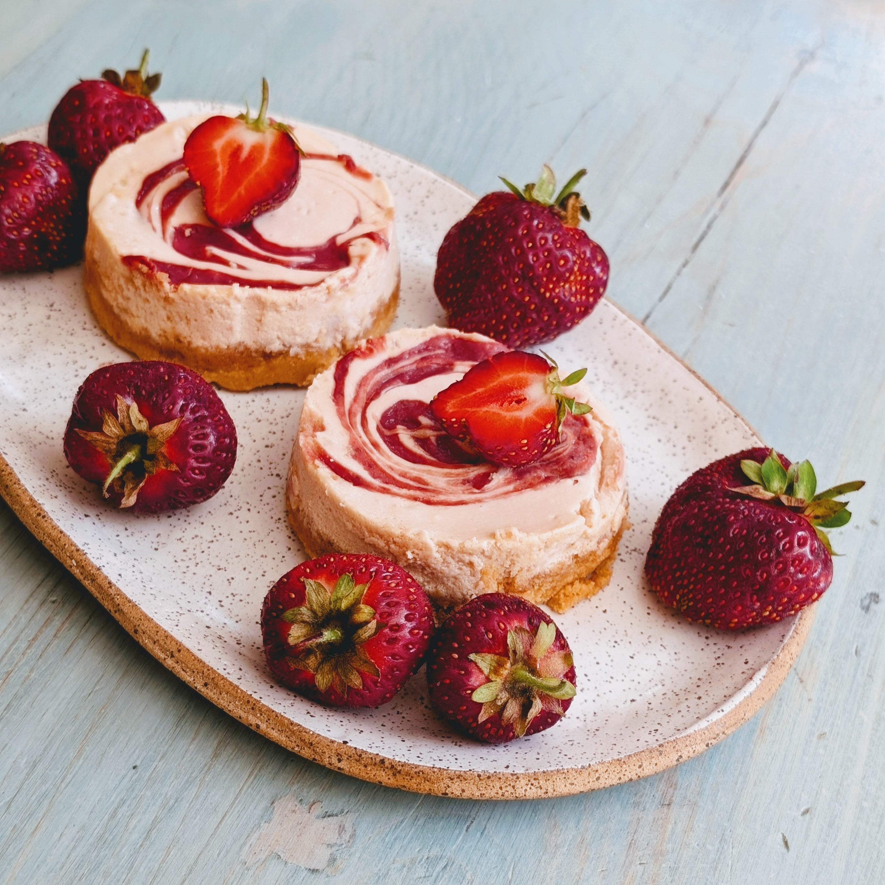 Two mini strawberry swirl cheesecakes on a speckled oval plate surrounded by fresh strawberries on a light blue table.