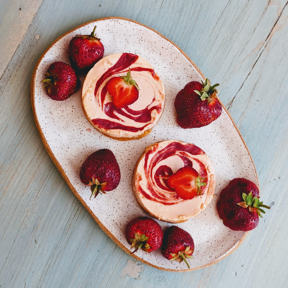 Two strawberry-swirl tarts on a speckled oval plate surrounded by whole strawberries on a light blue wooden table.