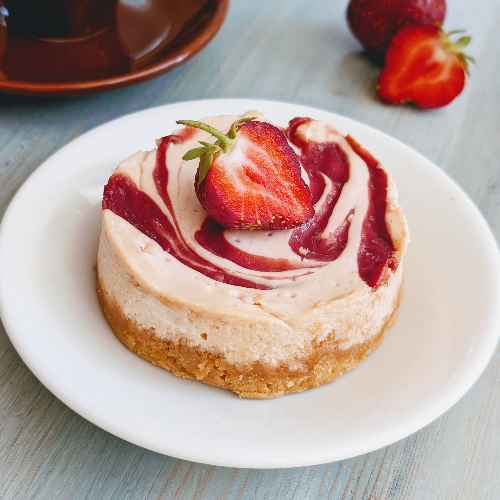 Mini strawberry swirl cheesecake with graham crust and halved strawberry on top, on a white plate