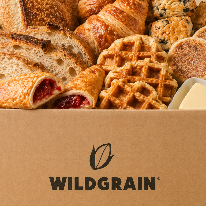 A Wildgrain box filled with assorted baked goods, including croissants, bread loaves, scones, English muffins, a waffle, and a small dish of butter, with the Wildgrain logo printed on the front against a warm beige background.