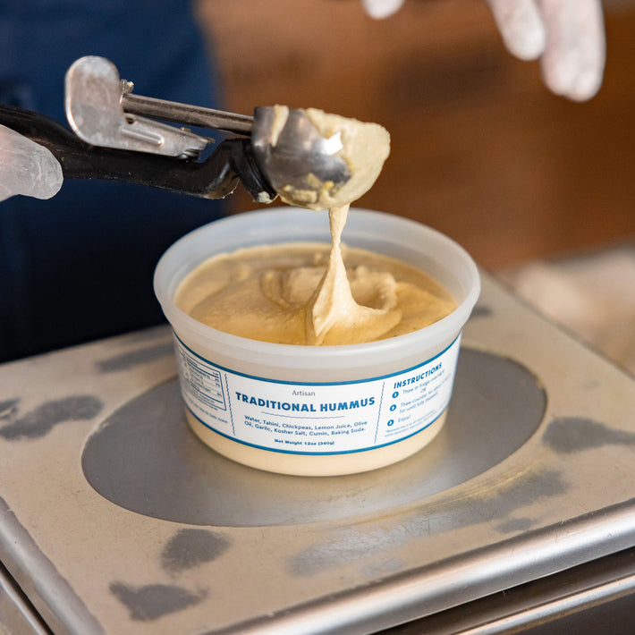 Person scooping hummus from a container with a spoon, wearing gloves.