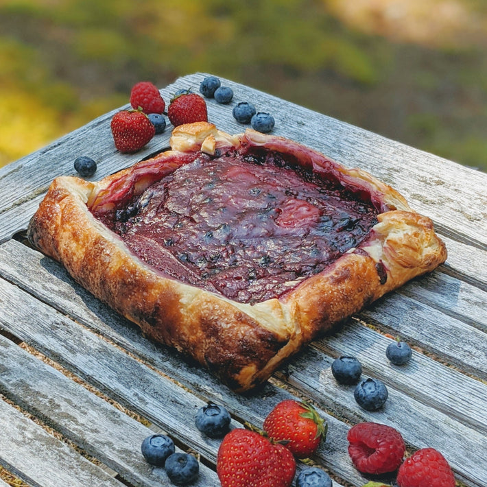 A rustic pastry filled with berry jam, surrounded by fresh raspberries and blueberries, on a wooden table.