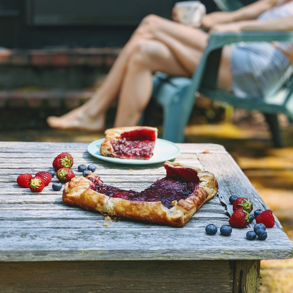 Mixed-berry galette missing a wedge on a weathered wooden table, strawberries and blueberries scattered, blurred seated person