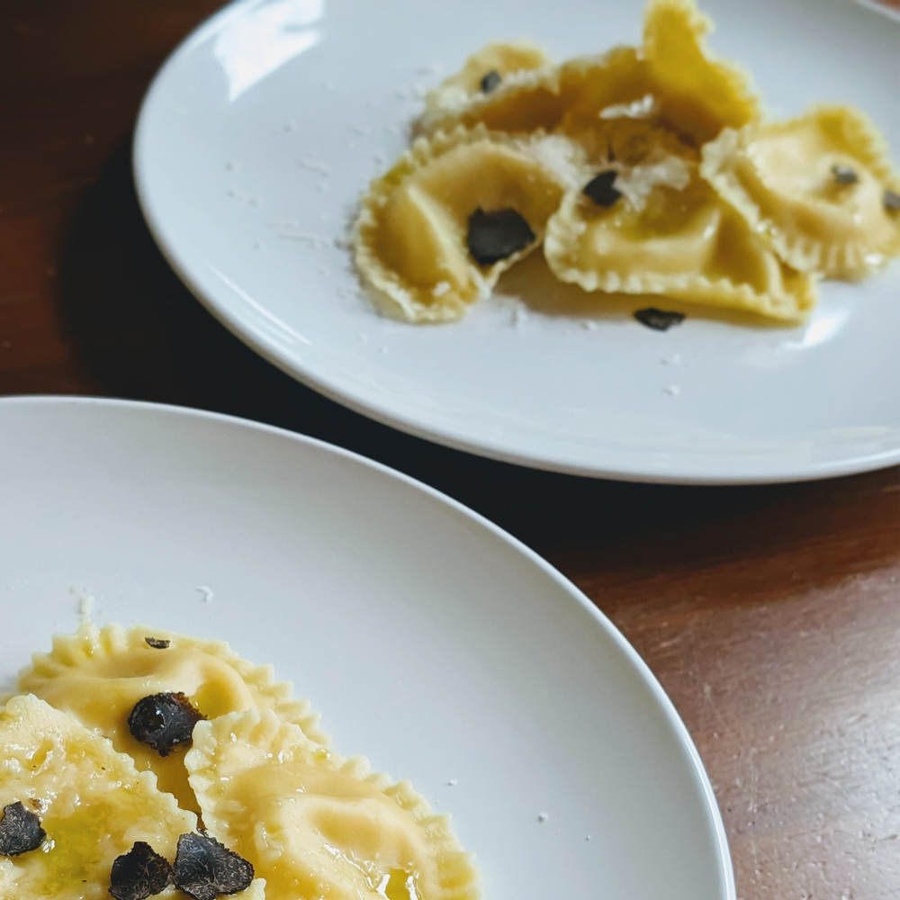 Two white plates of ravioli topped with shaved black truffle and grated cheese on a wooden table