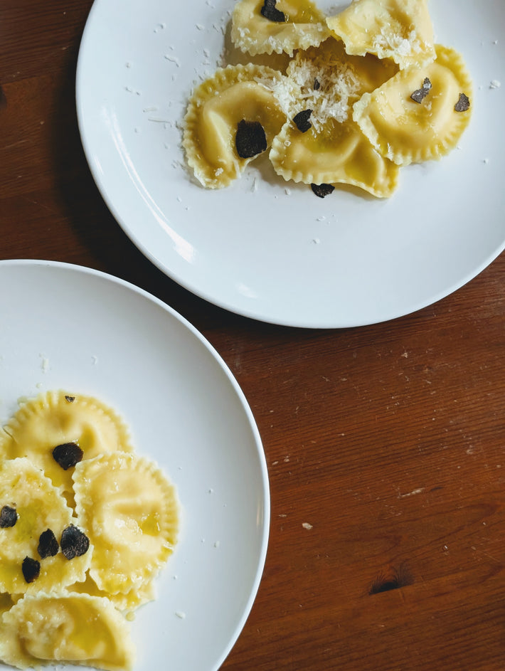 Two white plates on a wooden table, each with several pieces of ravioli topped with black truffle slices and grated cheese.