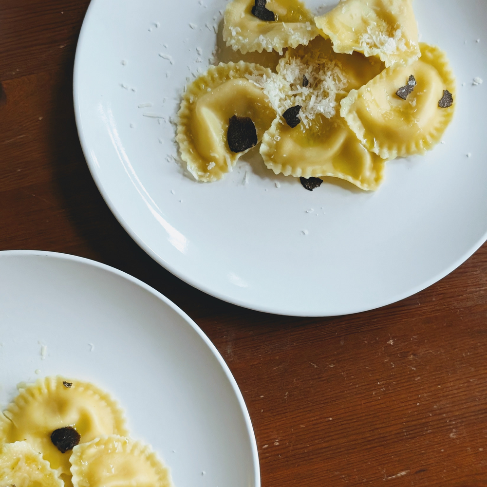 Ravioli on white plates, topped with grated cheese and slices of black truffle on a wooden table.