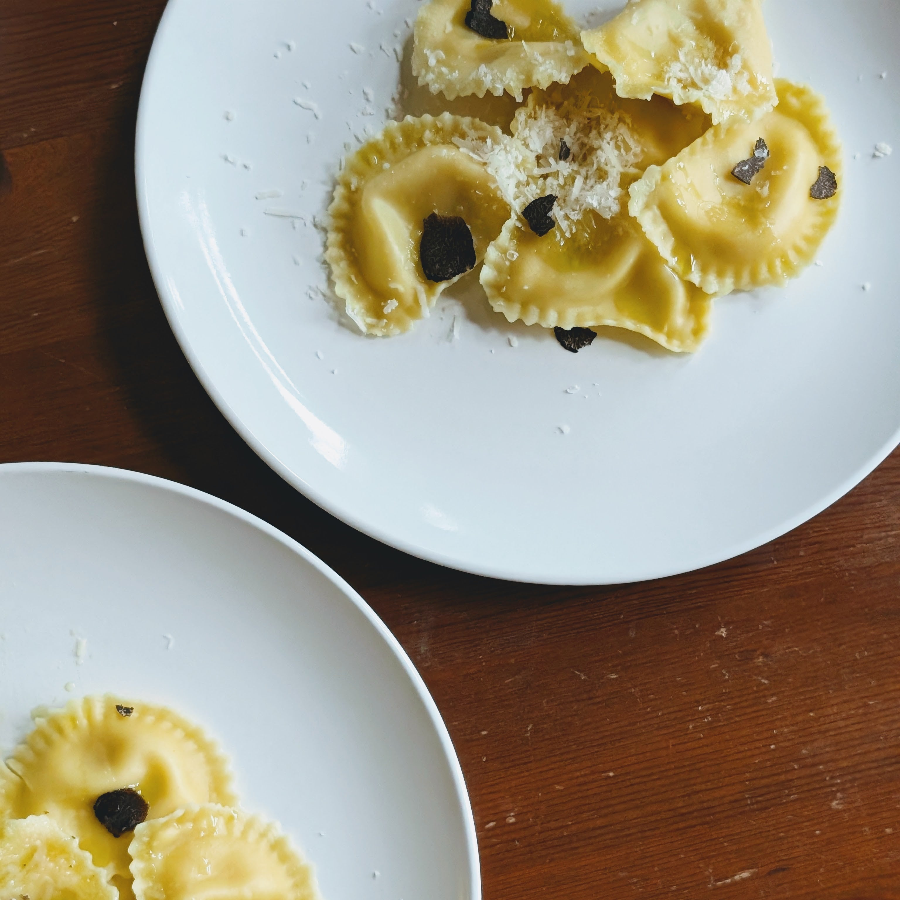 Ravioli on white plates, topped with grated cheese and slices of black truffle on a wooden table.
