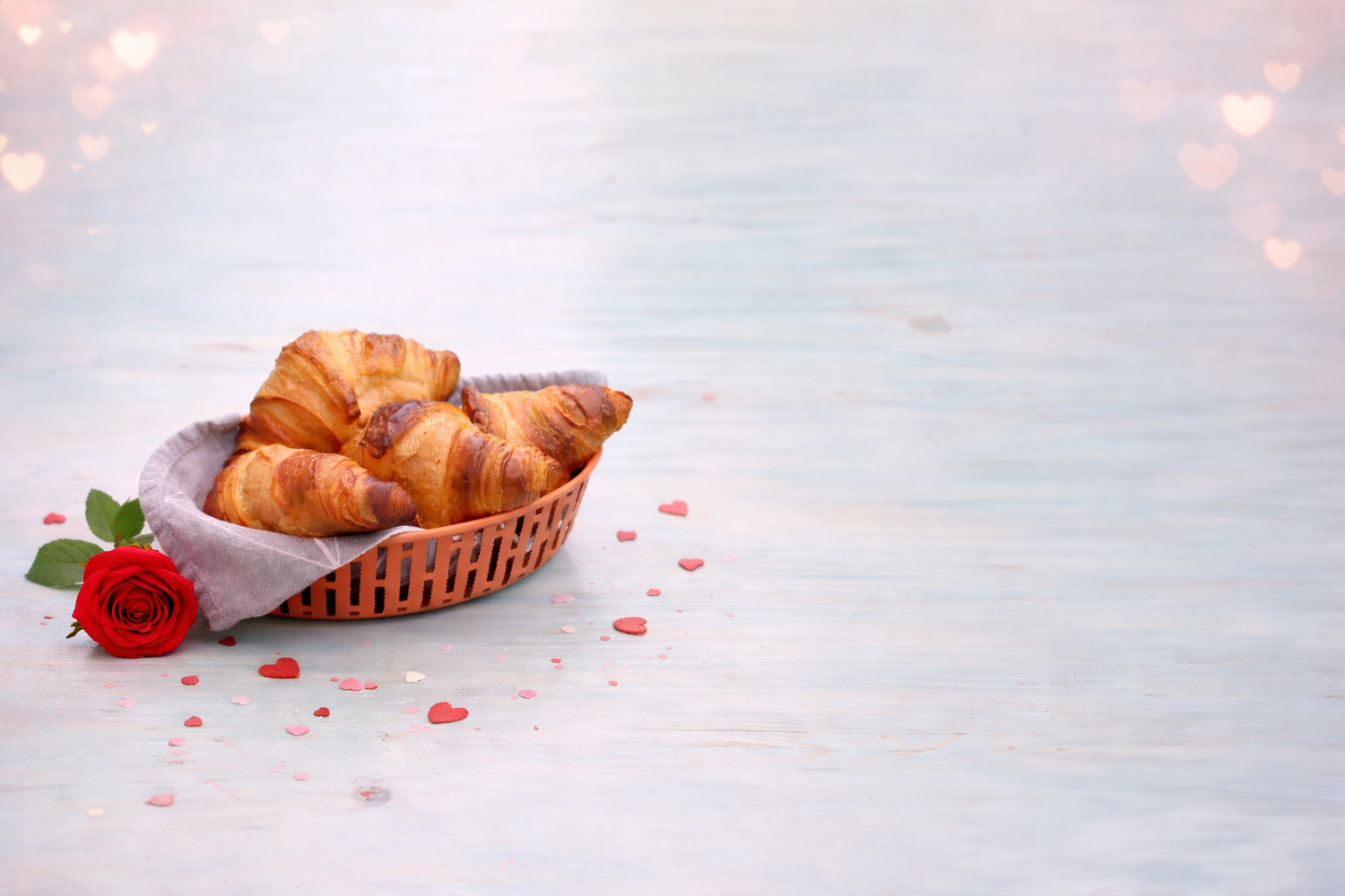 Basket of croissants with a red rose on a light wooden surface