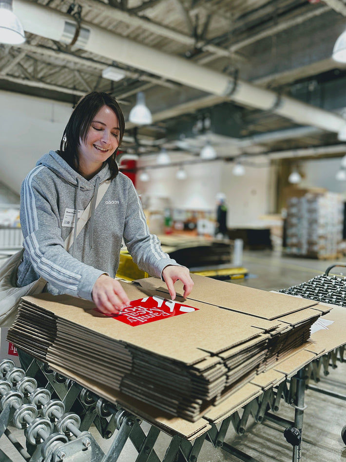 Smiling woman in gray adidas hoodie folding stacked cardboard boxes on a warehouse conveyor; red sticker reads 'Family Feast'.