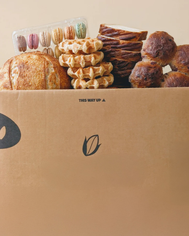 Cardboard box filled with bread, waffles, macarons and bagels; box label reads "THIS WAY UP".