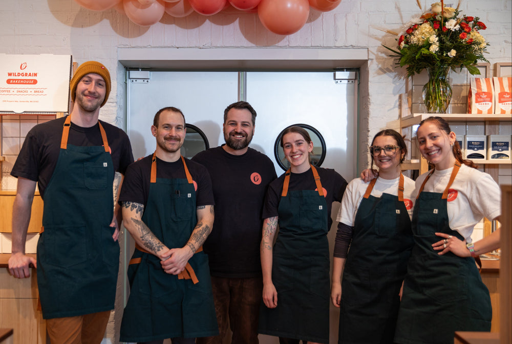 Team of people wearing aprons in a kitchen setting with balloons and flowers in the background.