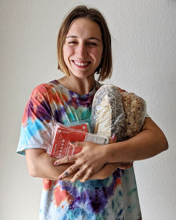 Smiling woman in tie-dye shirt holding two wrapped loaves of bread and packaged grocery items against a light wall.