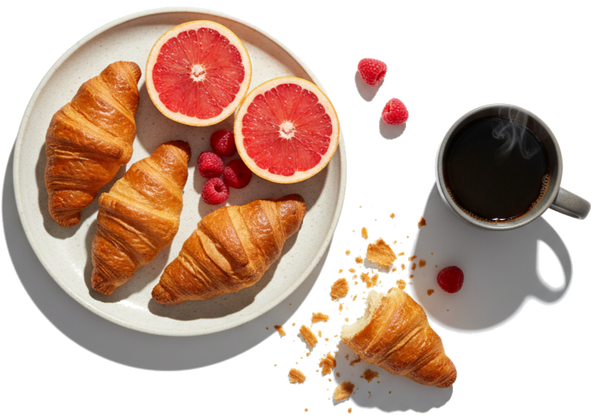 Three croissants with halved grapefruit and raspberries; a broken croissant and crumbs beside a steaming cup of coffee.
