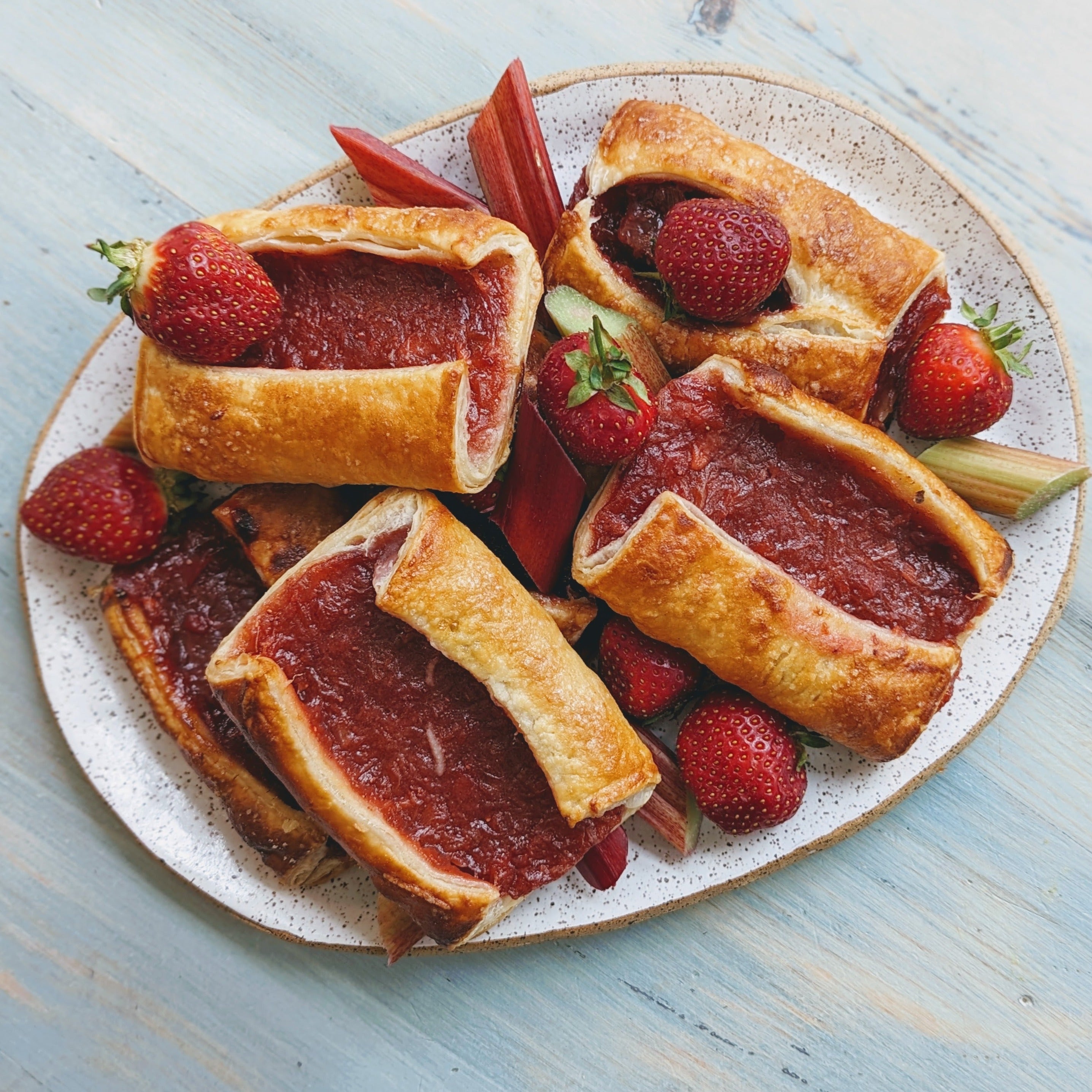 Plate of rectangular pastries filled with red fruit filling, garnished with fresh strawberries and rhubarb pieces on a light blue wooden surface.