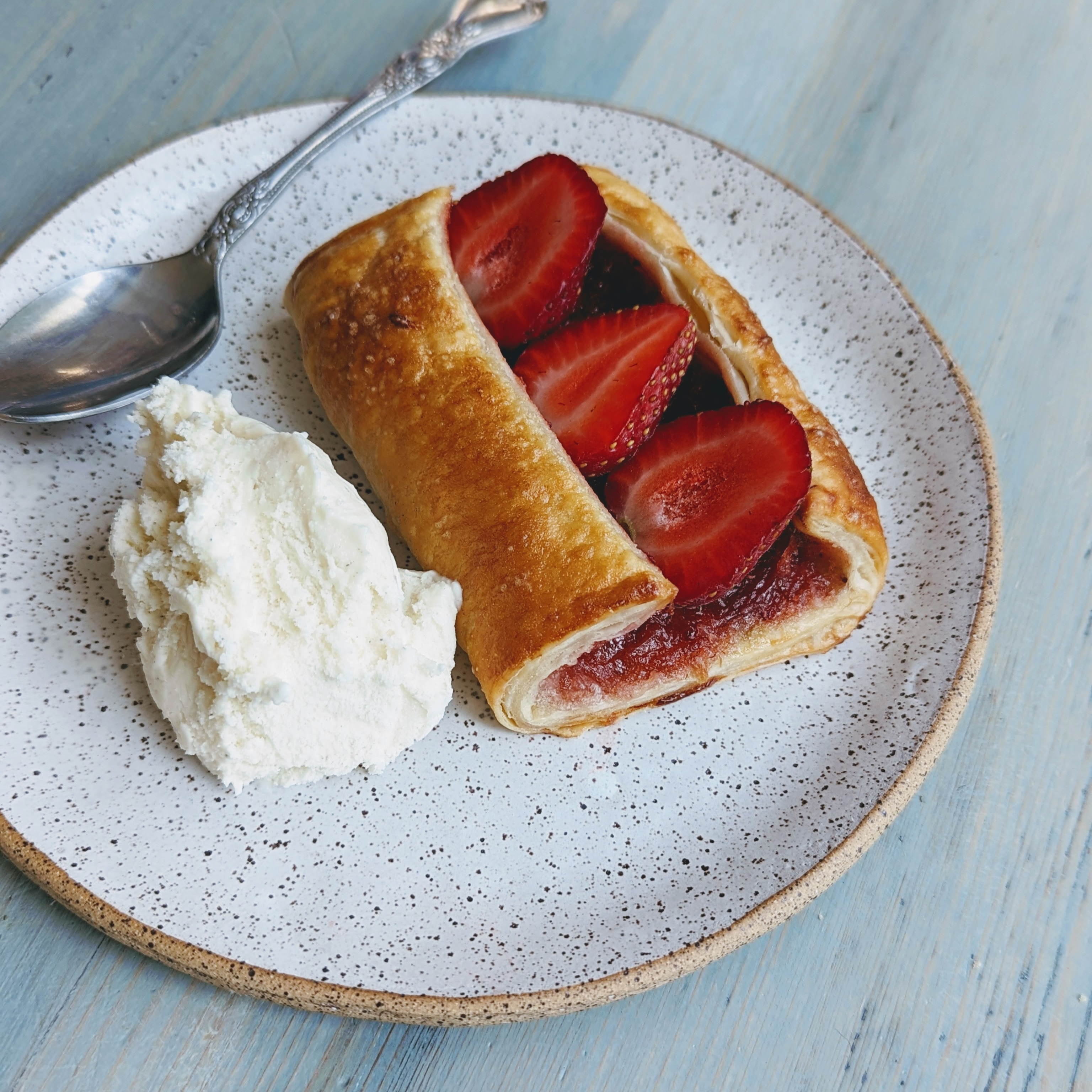 Strawberry-filled puff pastry with three sliced strawberries and a scoop of vanilla ice cream on a speckled plate with a spoon.