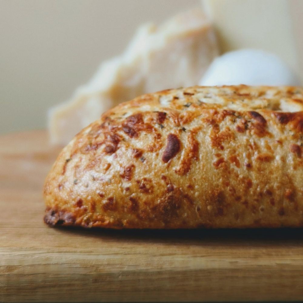 Golden cheese-topped bread roll on a wooden cutting board, with blurred cheese and an egg in the background.