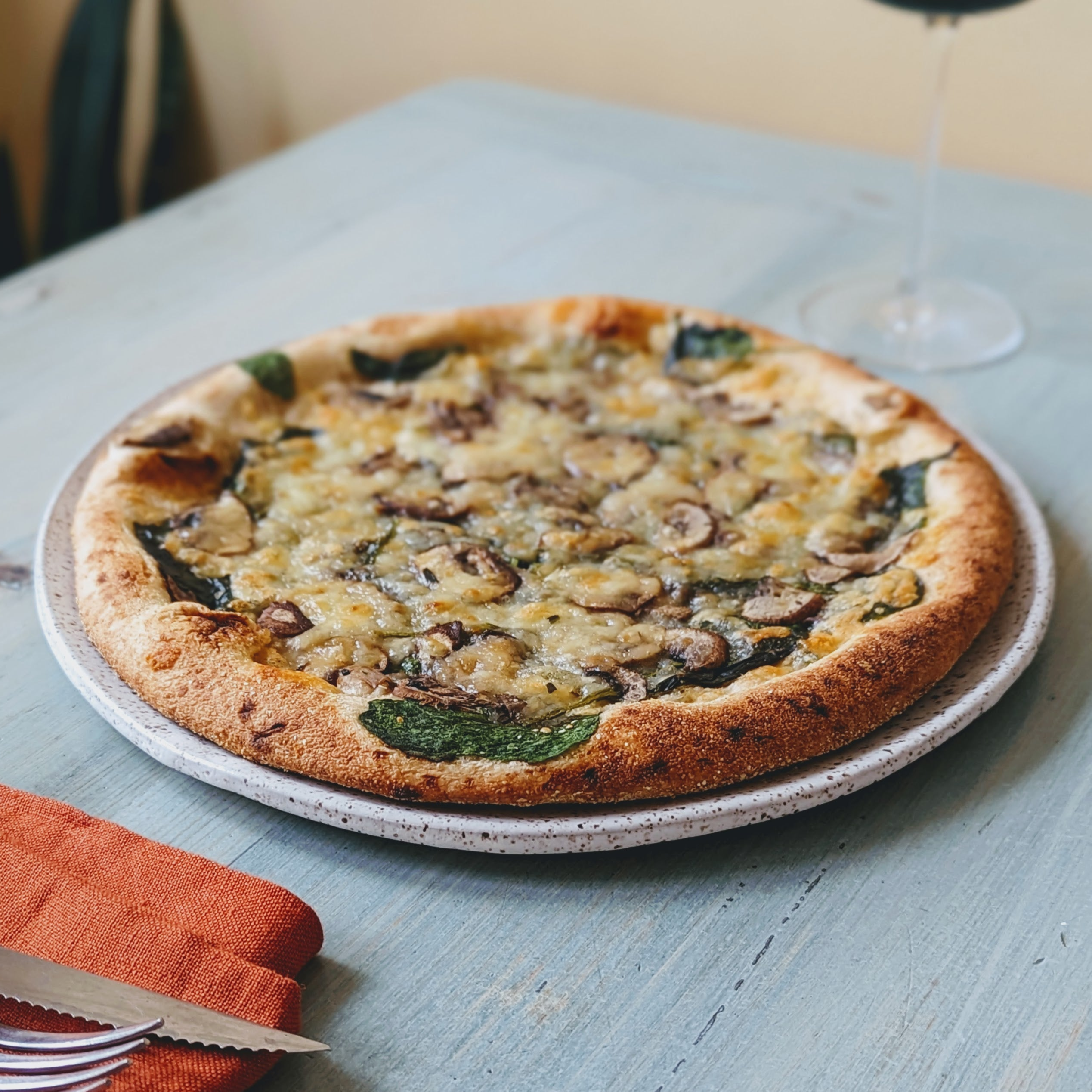 Mushroom and spinach pizza on a speckled ceramic plate on a light-blue wooden table with wine glass and orange napkin.