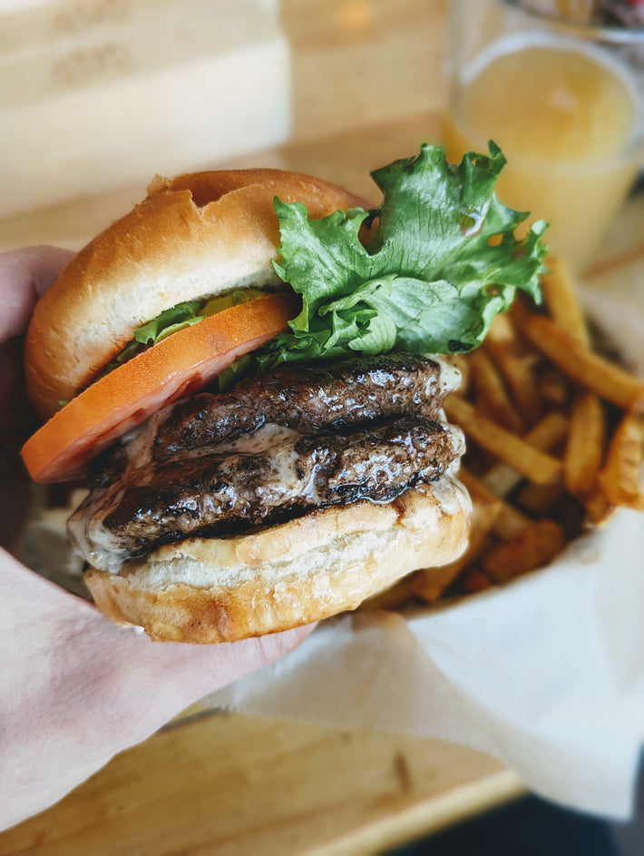A hand holding a hamburger with two beef patties, lettuce, tomato, and cheese, with fries in the background.