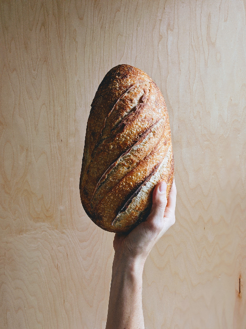 Hand holding a rustic oval loaf of crusty sourdough bread against a light wood background.
