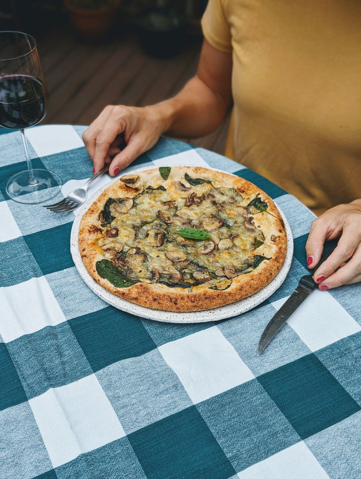 Hands cutting a mushroom-and-spinach pizza on a blue checkered tablecloth, wine glass at left.