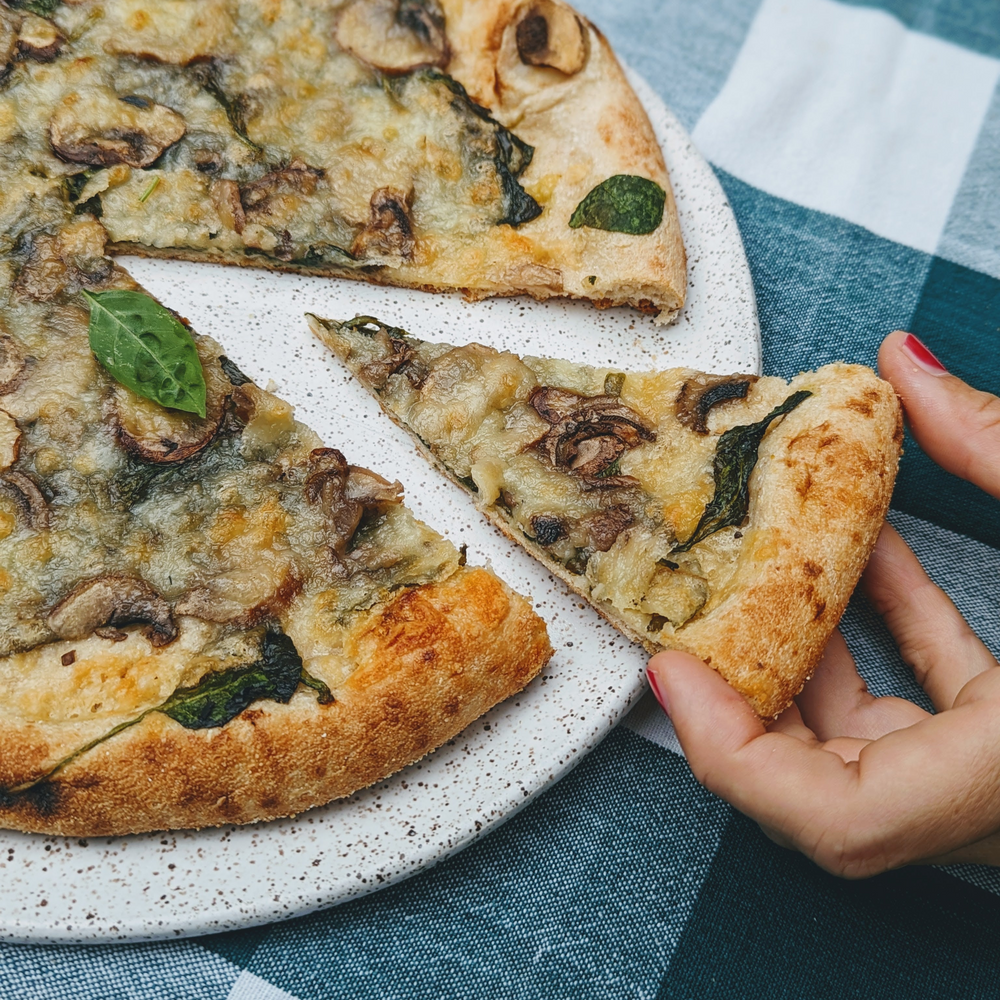 Hand picking up a slice of mushroom-and-spinach pizza from a speckled plate on a blue-checkered cloth.