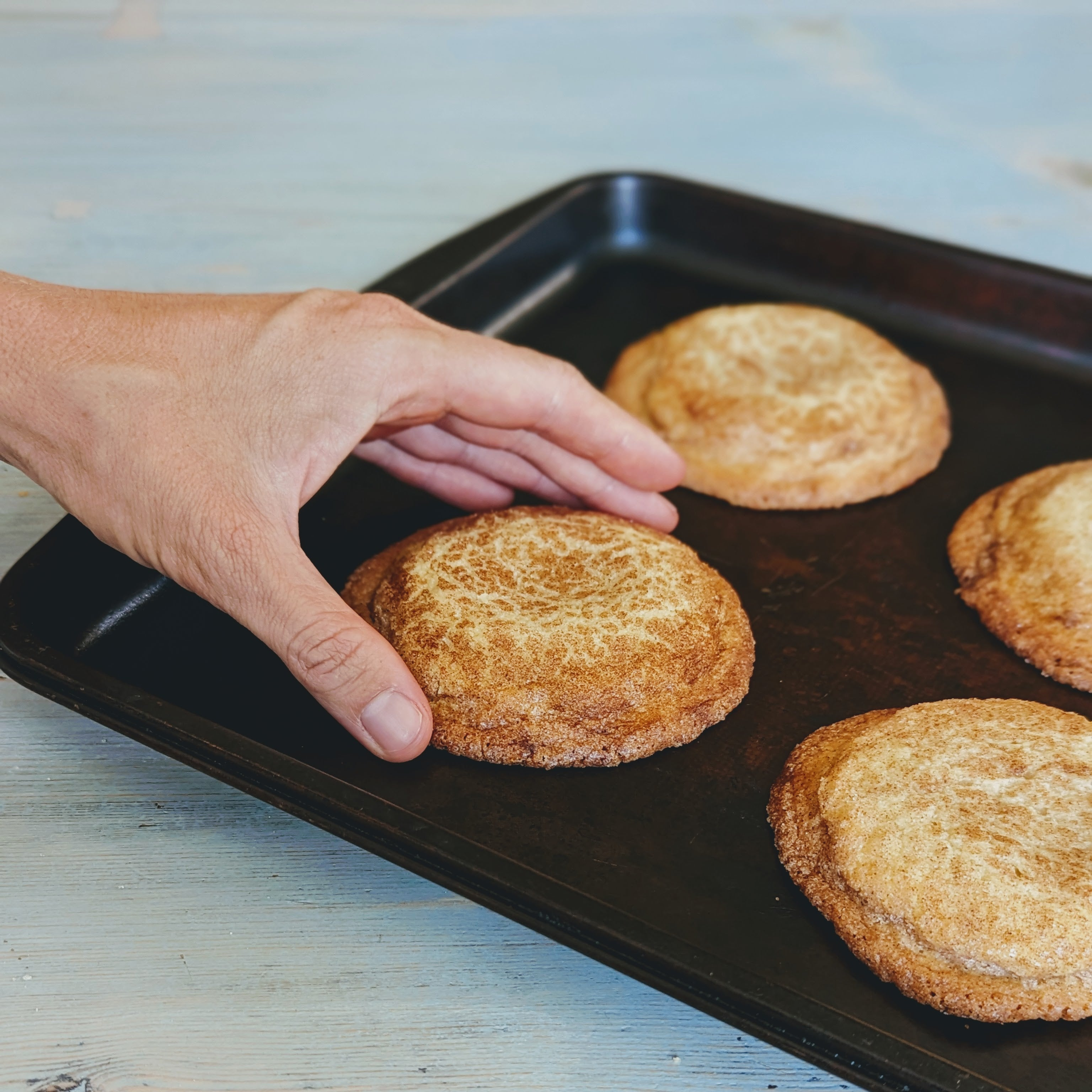 Hand reaching to pick a snickerdoodle-style cinnamon sugar cookie from a black baking sheet with three other cookies.