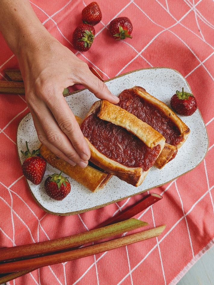 A hand holding a plate of Strawberry Rhubarb Turnovers with fresh strawberries and rhubarb on a pink tablecloth.