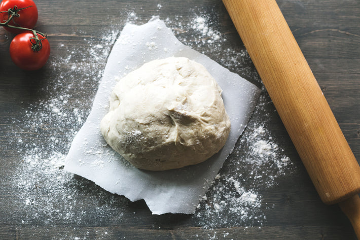 A ball of dough on parchment paper, surrounded by flour, with a rolling pin and cherry tomatoes nearby.